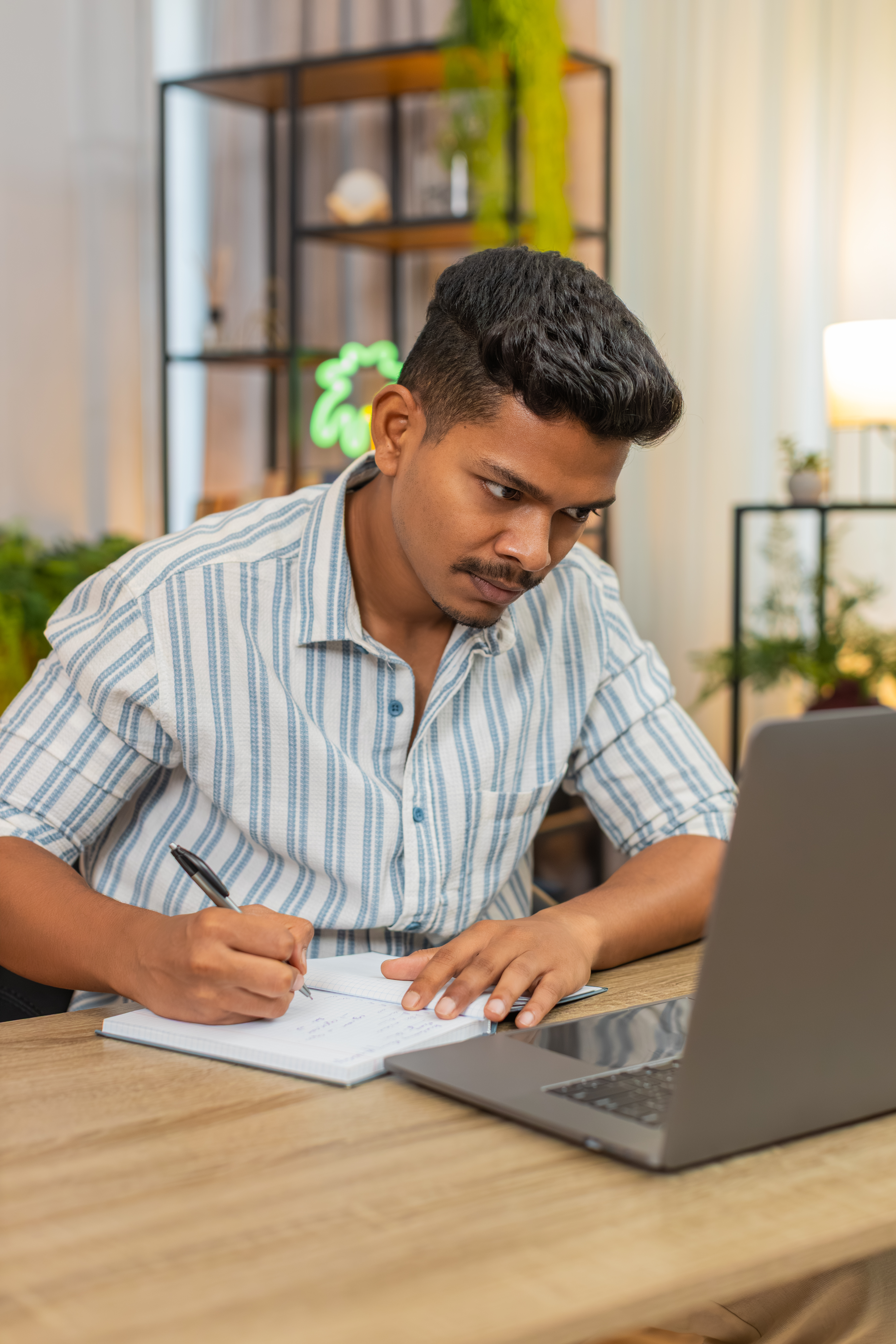 Indian man at home studies online course with laptop and notebook writes notes completes test. Arabian guy at table watches lecture reviews slides uses submits exam checks score and final grade.